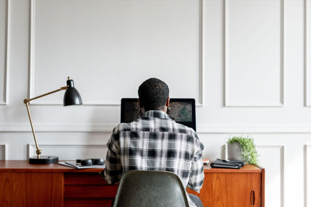 A man wearing a black and white flannel shirt faces a white paneled wall as he works at his laptop in his workplace.