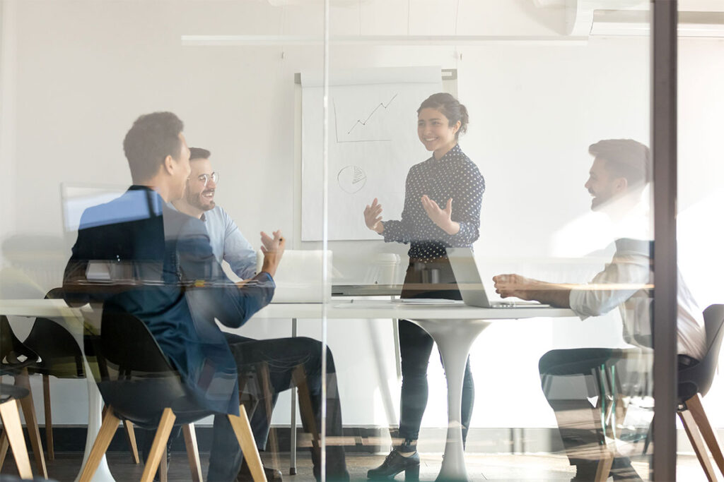 Four professionals work in a glass-walled conference room, with one team member standing and presenting.